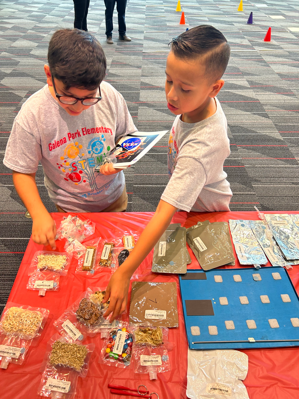 Two young boys look down at a display on an orange table. Both are wearing grey shirts with colored lettering.